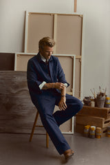 Wearing the BGliam Nebraska Chino in Blue by Blue de Gênes—a pair of high-density dobby weave cotton chino trousers—a person sits on a stool in an art studio, rolling up their sleeve amid large canvases, paintbrushes, and jars of paint.