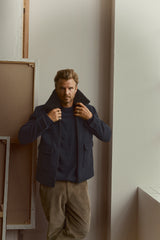 A man with styled hair and a beard stands indoors beside a window, leaning against a wall adorned with framed canvases. He is wearing the BGdugal Bomber Jacket in Dark Navy by Blue de Gênes, paired with corduroy pants and rings.