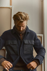 A man with tousled blond hair looks down while buttoning the Blue de Gênes BGBoyd Wool Jacket in Dark Navy over a brown turtleneck sweater, standing indoors in front of light-colored framed canvases.