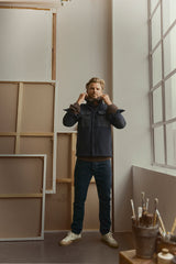 A man stands indoors near large canvases and a window, adjusting his jacket collar. He wears dark jeans, a brown sweater, white sneakers, and the BGBoyd Wool Jacket - Dark Navy by Blue de Gênes. A pot with paintbrushes sits on the floor nearby.