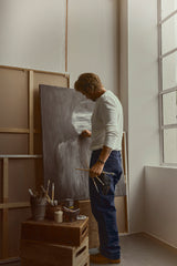 Wearing the BGliam Nebraska Chino in Blue by Blue de Gênes, a person dressed in a white long-sleeve shirt and equipped with an adjustable buckle strap paints on a canvas in their studio, holding brushes and standing near art supplies by a large window.
