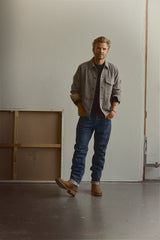 A man wearing the Blue de Gênes BGmungo Check Overshirt in Grey Check, paired with a dark shirt, jeans, and brown boots, stands indoors near a white wall and a framed canvas, looking at the camera with one foot slightly raised.