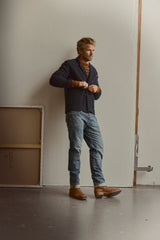 A man stands indoors near a wall, wearing a navy cardigan layered over the Blue de Gênes BGdaw Stripe Knit in Ginger Bread Orange, paired with blue jeans and brown boots. He is buttoning his cardigan while looking to the side.