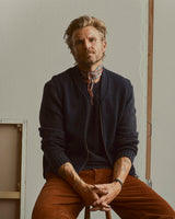A person with blond hair and a patterned scarf sits on a stool, wearing the Blue de Gênes BGadamo Crewneck Knit in Dark Navy and rust-colored pants, their tattooed forearms visible. The background is minimal, featuring a blank canvas.