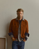 A man with light hair and a beard stands against a plain wall, wearing the Blue de Gênes BGhew Wool Overshirt in Ginger Bread Orange over a gray shirt and blue jeans, with his hands in his pockets.