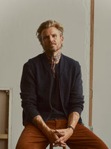 A man with blond hair and a beard sits on a stool against a plain wall, wearing a dark jacket layered over the Blue de Gênes BGmudock Wool Waistcoat in Dark Navy, paired with orange pants and a patterned scarf. His hands rest on his lap, revealing several tattoos.