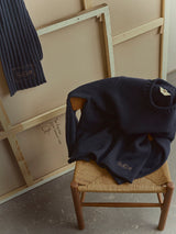 A navy blue sweater and the BGcomforto Knit Scarf – Dark Navy by Blue de Gênes, both accented with embroidered initials, rest on a woven chair beside the back of a large canvas leaning against a softly lit wall.