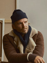 A bearded man wearing a brown shearling jacket, a navy knit scarf, and the BGboshi Beanie in Dark Navy by Blue de Gênes sits indoors, gazing thoughtfully to the side.