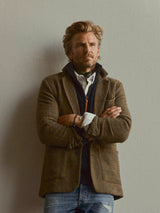 A man with wavy blond hair and a beard stands against a plain wall, arms crossed, wearing a brown blazer layered over the Blue de Gênes BGfraser Zip Knit in Dark Navy, paired with light blue ripped jeans.
