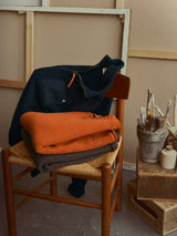 A wooden chair draped with the Blue de Gênes BGgordon Knit in Ginger Bread Orange, along with other folded wool sweaters—including a navy crew neck—is set against a neutral-toned background. Next to the chair, a stool holds paintbrushes in a container and various art supplies.