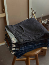 A neatly folded stack of Blue de Gênes BGleroy Jeans - Raw Denim sits on a wooden stool, with art supplies and canvases softly blurred in the background.