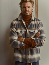 A man with light hair and a beard stands with arms crossed against a neutral wall, wearing a brown turtleneck sweater layered under the Blue de Gênes BGyodo Check Overshirt in Grey Check.