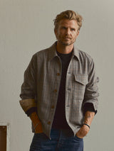 A man with light hair and a beard stands against a plain wall, wearing the Blue de Gênes BGmungo Check Overshirt in Grey Check over a black top and jeans, with one hand in his pocket.