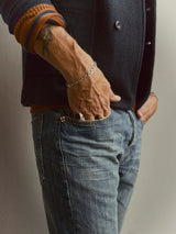 A person stands with one hand in their pocket, showing off a bracelet, ring, and a forearm tattoo, dressed in a dark jacket, orange sweater, and Blue de Gênes BGgiacomo Alfie Jeans in Mid Blue Denim.