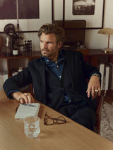 A man with styled hair, dressed in a dark three-piece suit and denim shirt, wears the Blue de Gênes BGduncan Ian Gilet in Black—a linen waistcoat crafted from lightweight textured fabric—as he sits at a wooden table with a notebook, glasses, and a glass of water. The background is adorned with framed art, books, and decorative objects.