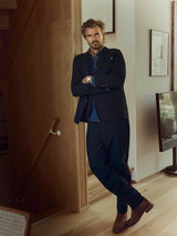 A man with a beard stands indoors, leaning against a wooden wall near the stairs. He wears the Blue de Gênes BGleopardi Ian Blazer in Black, paired with a blue shirt, brown boots, and a watch. With his arms crossed and looking at the camera, he is framed by a TV and framed art in the background.