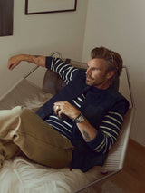 A man with light brown hair and tattoos relaxes on a hammock chair. He wears the Blue de Gênes BGdand Waistcoat in Dark Navy over a striped sweater and tan pants, looking thoughtfully to the side in a cozy, softly lit room.