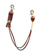 Introducing the BGsavino Key Hanger - Brown by Blue de Gênes: crafted from brown leather, this practical accessory features a double-ended design with brass clasps. It is adorned with a leather patch displaying a white cross on a red background and includes a wrapped section near one clasp. A sturdy logo rivet provides both durability and distinctive style.