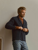 A man with light brown hair and a beard wears the BGlewis Wool Blazer in Dark Navy by Blue de Gênes over a brown striped sweater and jeans, standing against a plain light wall and gazing thoughtfully to the side.