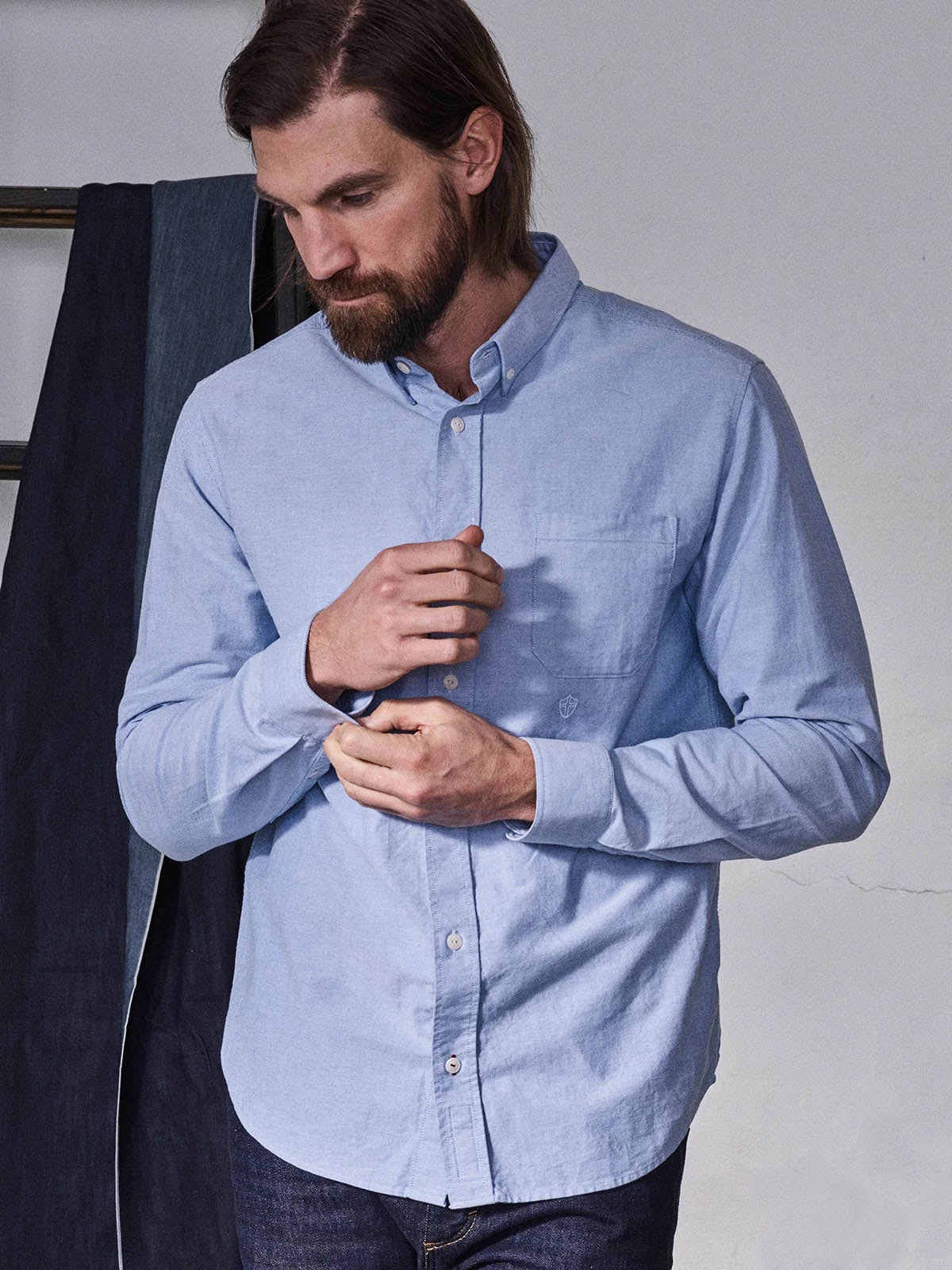 A man with long hair and a beard wears the Blue de Gênes BGsandro Oxford Shirt in blue paired with dark jeans, looking down as he adjusts his cuff. A dark fabric hangs in the background.