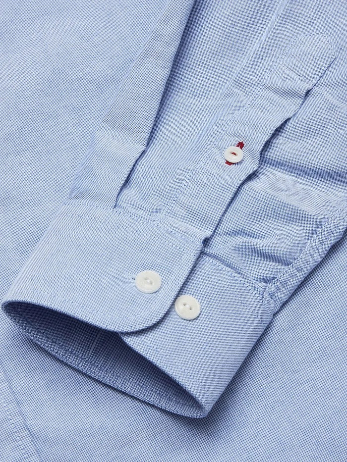 Close-up of the BGsandro Oxford Shirt - Blue by Blue de Gênes, featuring the sleeve cuff with two white buttons and part of the placket, which includes a small red-stitched buttonhole above another white button. The organic cotton fabric showcases a subtle textured pattern.