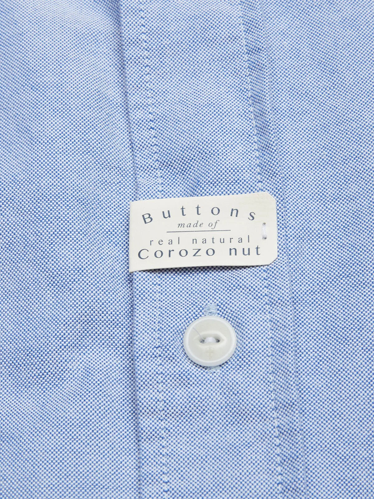 A close-up of the BGsandro Oxford Shirt - Blue by Blue de Gênes, made from light blue organic cotton, features a white label reading “Buttons made of real natural Corozo nut” above a single white button.