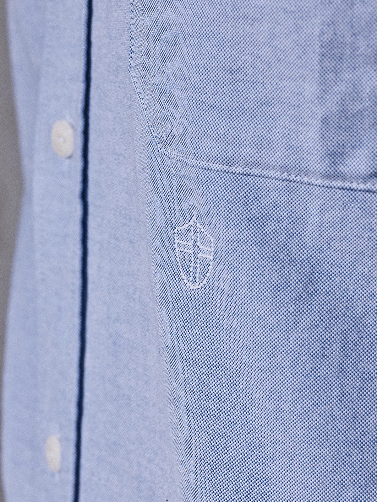 Close-up of the BGsandro Oxford Shirt - Blue by Blue de Gênes, featuring white buttons and a small white stitched shield emblem on the chest pocket.