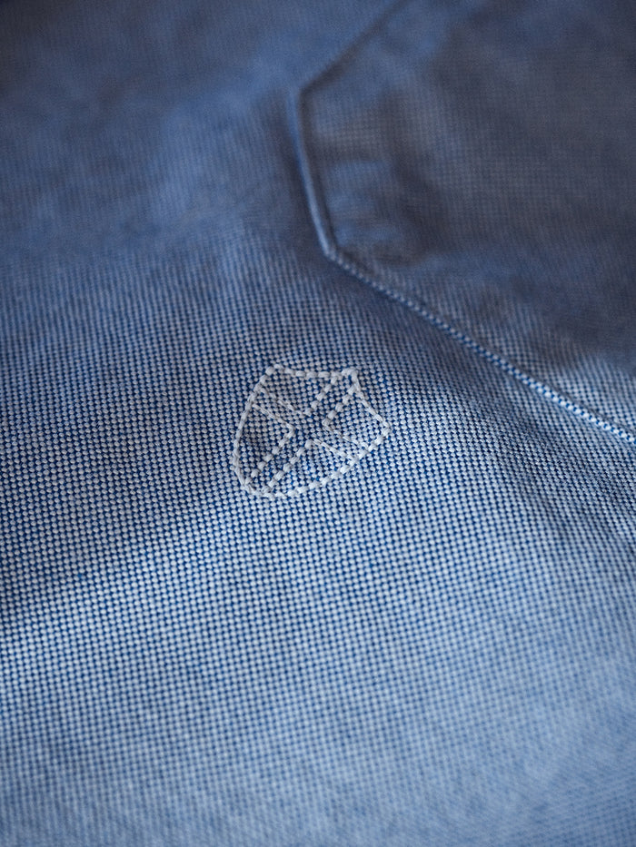 Close-up of the BGsandro Oxford Shirt - Blue by Blue de Gênes, crafted from organic cotton with a fine, textured weave and featuring a small white embroidered shield-shaped logo near the seam.