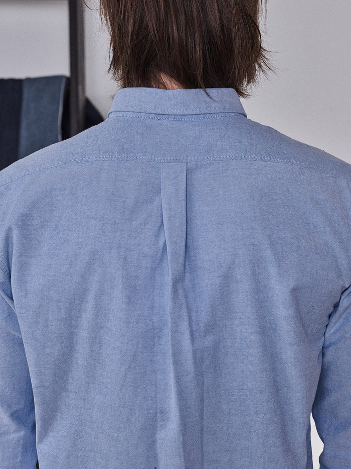 A person with shoulder-length brown hair is seen from behind, wearing the BGsandro Oxford Shirt - Blue by Blue de Gênes. The shirt features a center back pleat, and a dark garment hangs in the background.