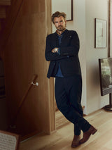 A man with a beard, dressed in a dark suit and Blue de Gênes BGpalco Ian Pant - Black, paired with boots, stands with his arms crossed near a wooden staircase in a modern room featuring light wood floors and minimal decor.