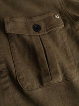 Close-up of the BGbarnaba Iro Overshirt in Beech Green by Blue de Gênes, highlighting its workwear-inspired olive green fabric, flap pocket with a black button, and a small metal shield-shaped pin on the top right corner of the flap.