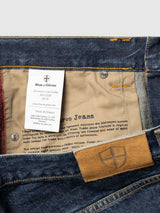 Close-up of BGmarco Cast Jeans - Denim wash by Blue de Gênes displays the inside label with product details, the Blue de Gênes logo, and a brown leather patch with a shield emblem on the waistband. The cotton denim fabric and stitching are clearly visible.
