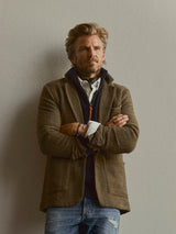 A man with light brown hair and a beard stands against a plain wall, arms crossed. He wears the Blue de Gênes BGenrico Tessusto Shirt in Chalk beneath a brown blazer and navy sweater, paired with slightly distressed blue jeans.