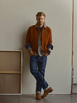 A man with light hair and a beard stands indoors against a plain wall, wearing a rust-colored jacket layered over shirts, Brown boots, gloves, and relaxed loose fit BGgiacomo Arthour Jeans in Denim Blue by Blue de Gênes, with two canvases leaning against the wall nearby.