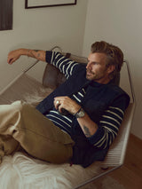 A man with light brown hair reclines on a hammock chair indoors, wearing the BGdaw Stripe Knit - Navy sweater by Blue de Gênes—a maritime-inspired, 100% wool striped crewneck—paired with tan pants and a dark vest. Tattoos adorn his left arm and wrist as he gazes thoughtfully to the side.