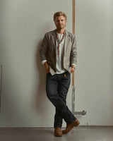 A man with blond hair and a beard leans against a wall, wearing a gray blazer, white shirt, scarf, and relaxed-fit BGmarco Connor Jeans in Black Denim Used by Blue de Gênes. His hands are in his pockets and he looks relaxed, effortlessly embodying style in 100% cotton denim.