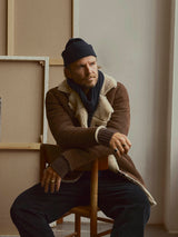 A man wearing the BGseras Shearling Coat in dark brown by Blue de Gênes, paired with dark pants, a knit beanie, and a scarf sits on a wooden chair in a neutral-toned room with canvases propped against the wall behind him.