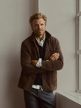 A man with blond hair and a beard stands indoors by a window, arms crossed. He wears a brown jacket, white shirt, and the Blue de Gênes BGmurry Polo Knit in Dark Navy beneath a scarf around his neck, looking confidently at the camera.