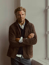 A man with blond hair and a beard stands by a window, arms crossed, wearing the BGvalan Suede Jacket in Dark Brown by Blue de Gênes over a sweater and collared shirt, and a patterned scarf around his neck.