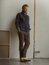 A man with light hair and a beard stands indoors against a pale wall, wearing a dark jacket, Blue de Gênes BGpaulo Ottawa V3 Trousers in Hot Fudge Brown, and beige boots. He has his hands in his pockets and looks down with a thoughtful expression.