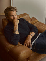 Man wearing a navy blue BGstu Rollneck Knit - Navy with a textured roll-neck, lounging in a leather armchair with his hand to his chin.