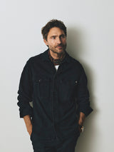 A man wears an enzyme washed, dark blue button-down shirt with a patterned neckerchief, his hands resting in the pockets of his BGpaulo Pavia V3 Chino - Midnight Blue trousers.