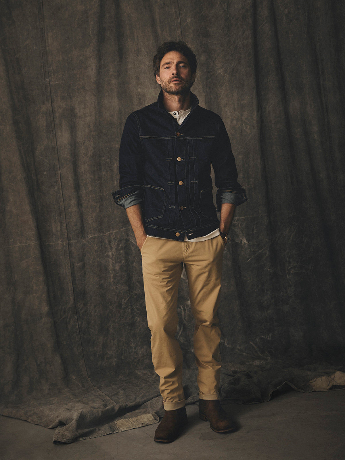A man wears a dark denim jacket over a white shirt and Blue de Gênes BGmenzo Vecchio Chino - Pale Khaki pants with brown boots.