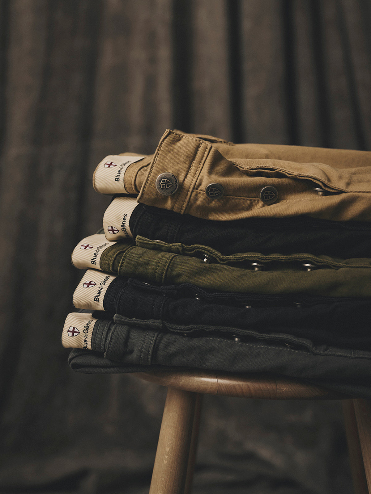 A stack of enzyme washed vintage military BGpaulo Pavia V3 Chino - Khaki in khaki, olive, black, and dark gray folded on a wooden stool.