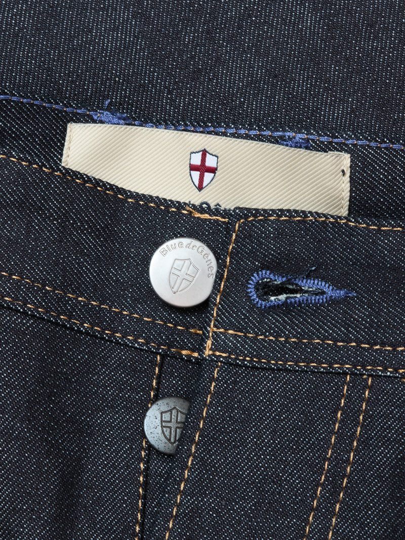 Close-up of Giacomo Selvedge V2 Jeans - Raw Denim by Blue de Gênes, featuring rigid denim, engraved metal buttons with a shield, gold stitching, and a beige label with a red and white cross. Relaxed loose fit. Made in Italy.
