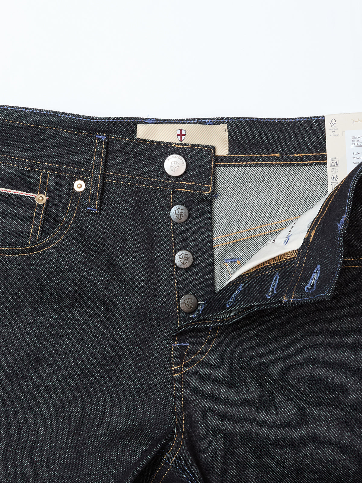 Close-up of Blue de Gênes Giacomo Selvedge V2 Jeans in raw denim: dark blue, rigid, relaxed loose fit with a partially open fly showing four metal buttons, inner lining, yellow stitching, and a white waistband patch with red emblem.