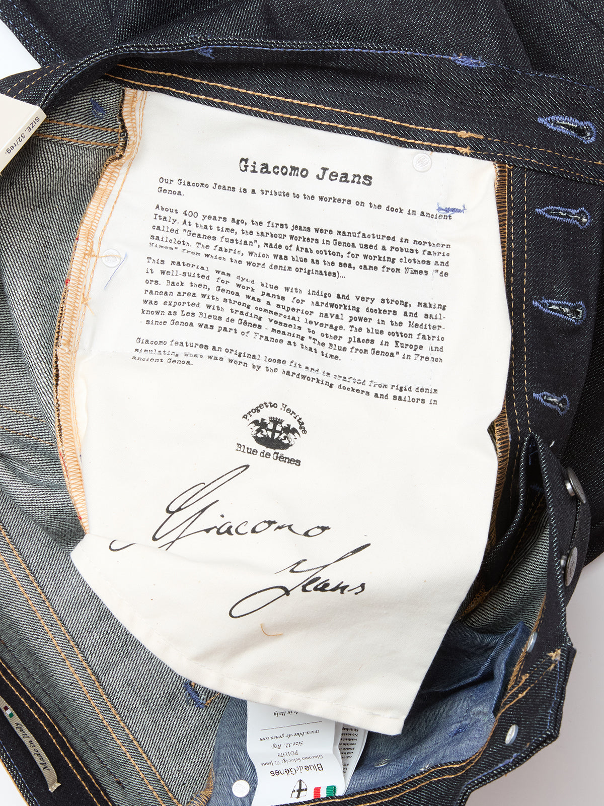 A close-up of the Giacomo Selvedge V2 Jeans - Raw Denim by Blue de Gênes, made in Italy, showing a printed label with brand story, logo, and signature on white fabric sewn inside the denim waistband and pocket area.