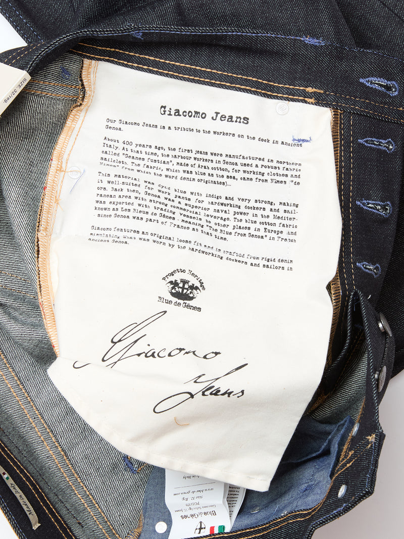 A close-up of the Giacomo Selvedge V2 Jeans - Raw Denim by Blue de Gênes, made in Italy, showing a printed label with brand story, logo, and signature on white fabric sewn inside the denim waistband and pocket area.