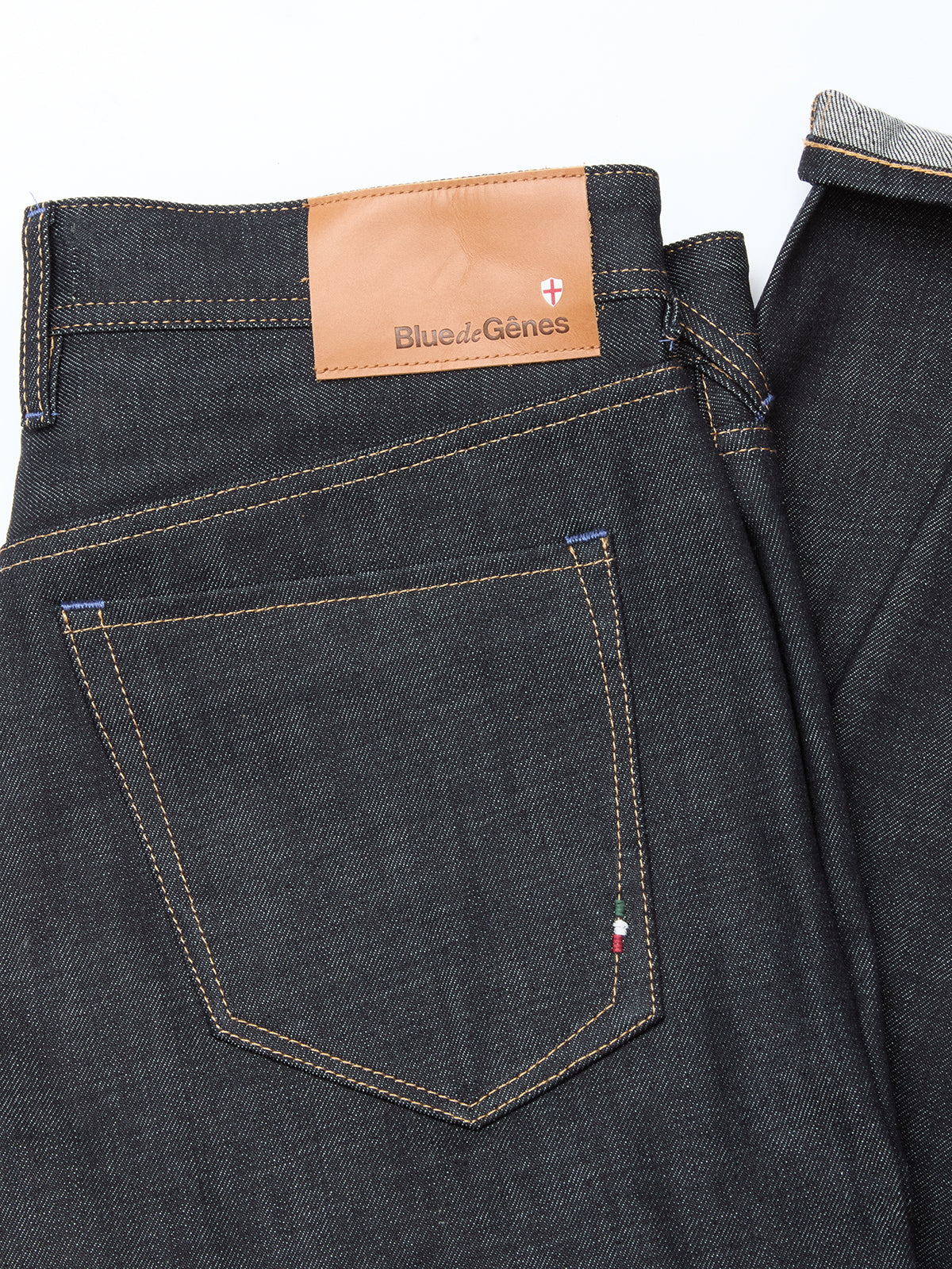 A close-up of the Giacomo Selvedge V2 Jeans - Raw Denim by Blue de Gênes, expertly made in Italy, shows dark blue rigid denim with brown stitching, a branded leather patch above the back pocket, and an embroidered Italian flag on the pocket edge.