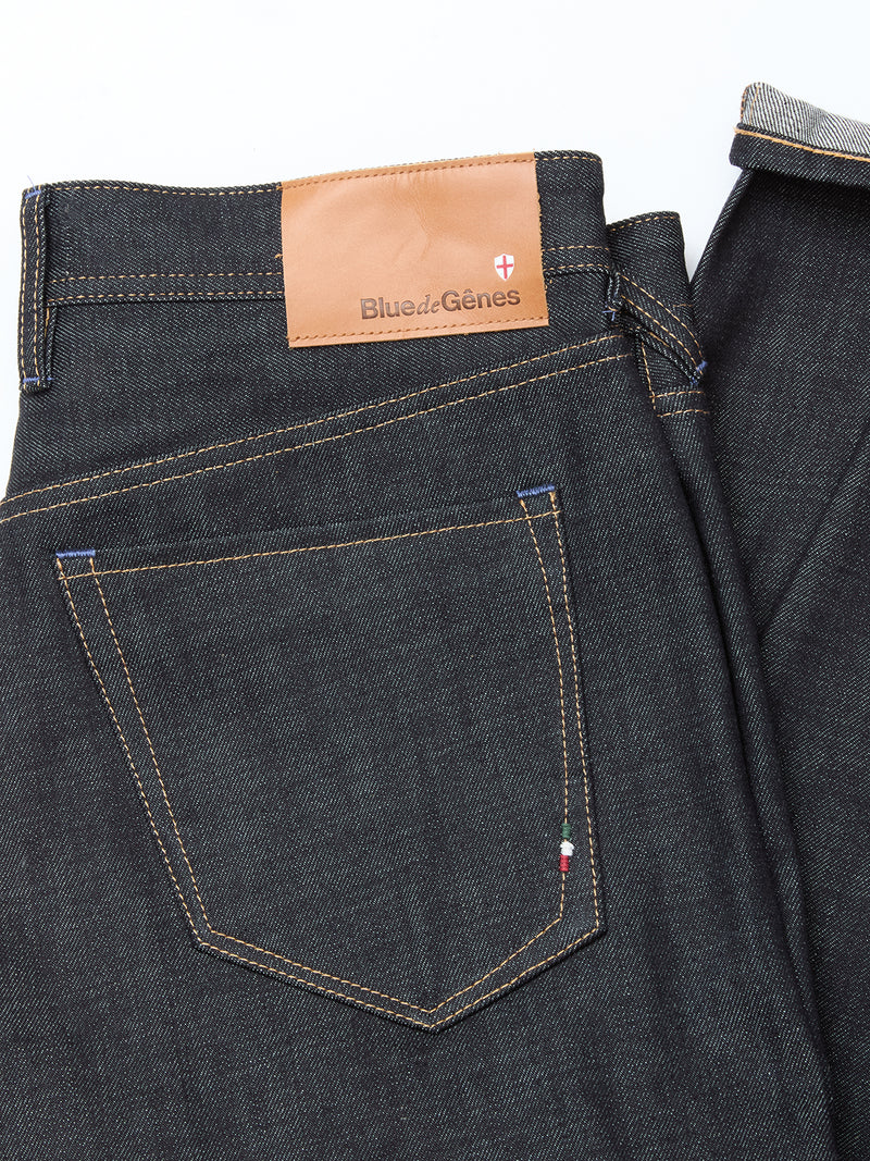 A close-up of the Giacomo Selvedge V2 Jeans - Raw Denim by Blue de Gênes, expertly made in Italy, shows dark blue rigid denim with brown stitching, a branded leather patch above the back pocket, and an embroidered Italian flag on the pocket edge.